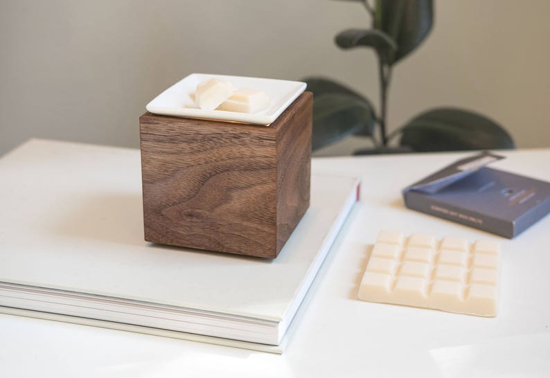 May include: A wooden cube diffuser with a white ceramic dish on top. The dish contains wax melts. The diffuser is sitting on a white book.