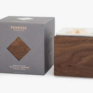 May include: A gray box with the text "Penrose Candles" and "Premium Solid Wood Wax & Oil Warmer" on it. The box is next to a wooden wax warmer with a white ceramic dish on top.