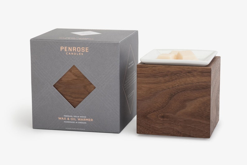 May include: A gray box with the text "Penrose Candles" and "Premium Solid Wood Wax & Oil Warmer" on it. The box is next to a wooden wax warmer with a white ceramic dish on top.