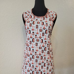 May include: A white apron with a red and black chess piece pattern. The apron has two pockets and a red and black trim.