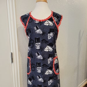 May include: A dark gray apron featuring a repeating pattern of skulls wearing top hats and spiders. The apron has red trim with white polka dots and two front pockets. The apron is displayed on a mannequin.