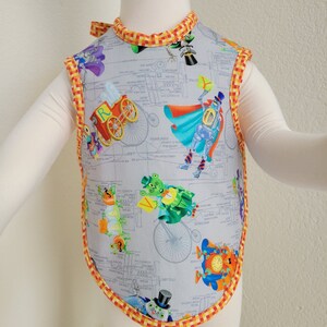 May include: A gray sleeveless bib with a colorful print of robots, trains, and other whimsical characters. The bib has a yellow and orange checkered trim.