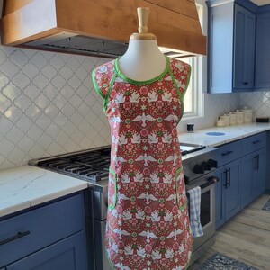May include: A red and white apron with a green trim and a pattern of white doves and flowers. The apron has a large pocket on the front and is displayed on a mannequin in a kitchen setting.