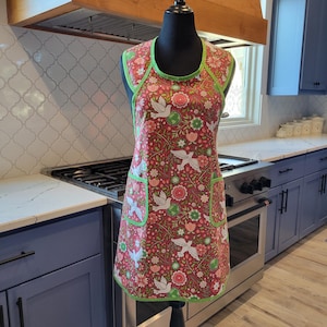 May include: A red apron with a floral and bird pattern, accented with green trim. The apron features two pockets and is displayed on a black mannequin. The kitchen setting includes a stainless steel stove and blue cabinets.