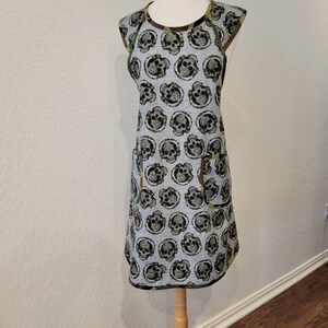 May include: A gray and green apron with a skull pattern. The apron has a large pocket on the front and a scoop neck.