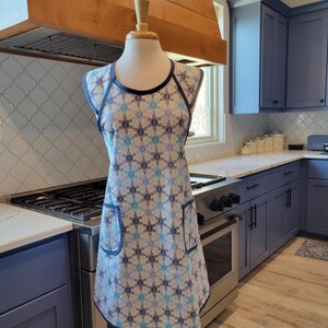 May include: A blue and white patterned apron with a navy blue trim and two pockets. The apron is on a white mannequin in a kitchen setting.