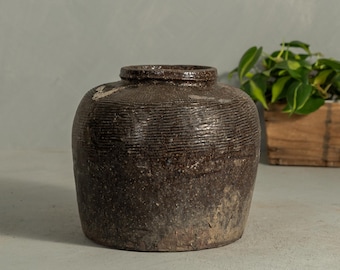 May include: A brown, textured ceramic jar with a wide base and a narrow neck. The jar has horizontal ridges and a slightly uneven surface. A small wooden box with green plants is in the background.