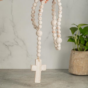 May include: A distressed white wooden rosary with large, round beads and a cross pendant. The beads vary in size and show hints of the wood grain. The rosary is held up against a marble-patterned background.