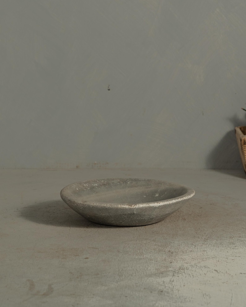 May include: A small, round, gray ceramic bowl with a textured surface. The bowl has a shallow depth and a slightly raised rim. The bowl is sitting on a gray surface.