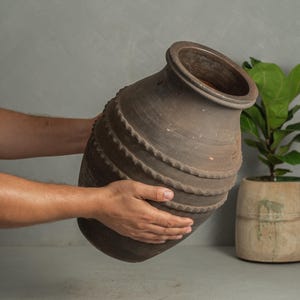 May include: A large, dark brown, matte-finished ceramic vessel with a wide, open mouth and a rounded body. The exterior surface features three distinct horizontal bands of raised, scalloped, or rope-like decorative texture encircling the middle.