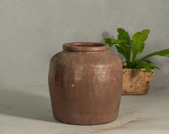 Op de afbeelding: Een brede, bruine keramische pot met een korte, dikke hals en een afgerond lichaam. Het bovenste gedeelte van de pot heeft een reeks verticale, ingesneden lijnen en horizontale banden, terwijl het onderste deel glad is.