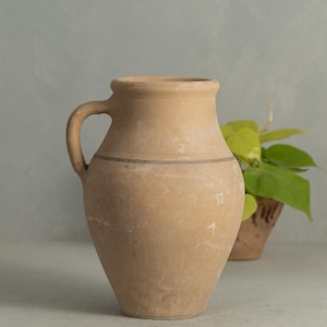 May include: A terracotta pitcher with a wide mouth and a single handle. The pitcher has a brown glaze and a dark brown stripe around the middle.