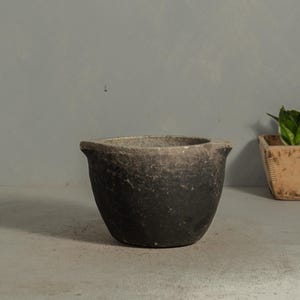 May include: A small, dark gray ceramic bowl with a wide opening and a slightly textured surface. The bowl has a rounded shape and a dark, almost black, color at the base, fading to a lighter gray at the rim. The background is a muted gray.