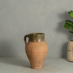 May include: A terracotta pitcher with a bulbous body and a single handle. The upper portion of the vessel features a textured, mottled olive-green glaze, while the lower section remains in a natural, matte, unglazed clay finish with visible surface variations.