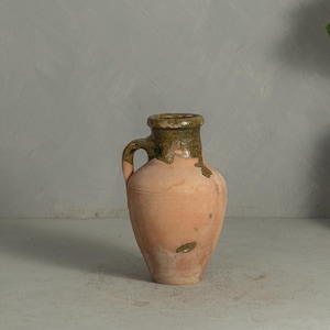 May include: A terracotta pitcher with a single handle and a wide, flared rim. The upper portion of the vessel features a mottled, dark olive-green glaze that drips down onto the matte, light peach-colored clay body, which shows natural wear.