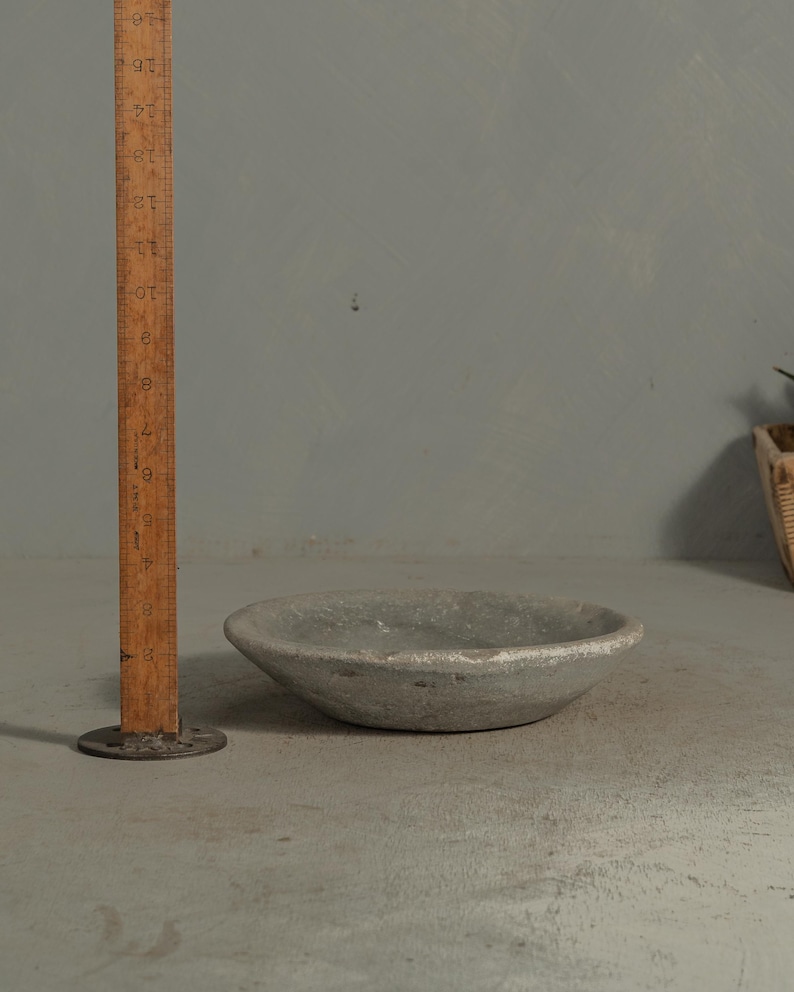 May include: A small, round, gray bowl with a rough texture. The bowl is approximately 6 inches in diameter. A wooden ruler stands next to the bowl, providing a size reference. The bowl sits on a gray surface.