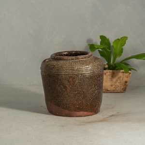 May include: A cylindrical ceramic jar with a wide mouth and a thick, rolled rim. The upper portion features a textured, ribbed pattern, while the lower body has a mottled, dark brown glaze with visible reddish-brown clay at the base.