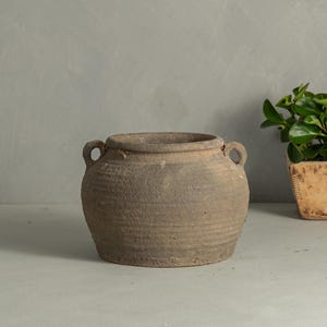 May include: A round, gray, textured clay pot with two small side handles. The vessel features a wide opening and a weathered, rustic surface with horizontal ridges around the body, showcasing a natural, earthy appearance and a sturdy, wide base.