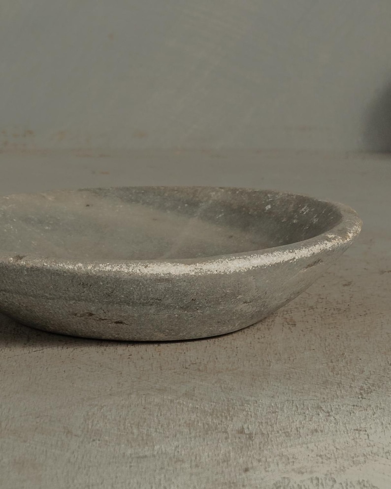 May include: A small, round, gray stone bowl with a slightly textured surface. The rim of the bowl has a white painted edge. The bowl is likely used for decorative purposes or for holding small items.