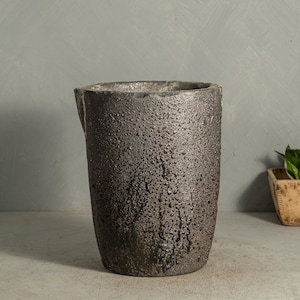 May include: A dark brown, textured ceramic vase with a cylindrical shape and a slightly flared top. The vase has a rough, pitted surface, and a small spout on the side. The vase is suitable for home decor.
