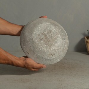 May include: A round, gray stone bowl with a textured surface. The bowl is held by two hands, showcasing its shape and material. The bowl's design suggests it could be used for decorative or functional purposes.