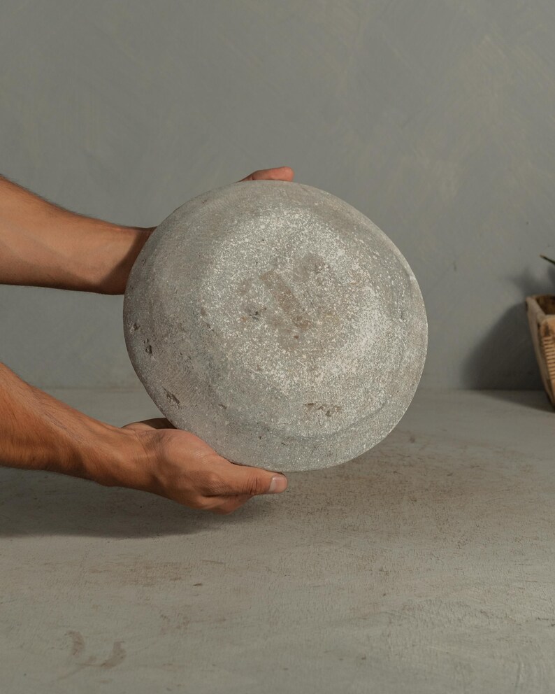May include: A round, gray stone bowl with a textured surface. The bowl is held by two hands, showcasing its shape and material. The bowl's design suggests it could be used for decorative or functional purposes.