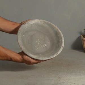 May include: A gray, round, stone bowl with a textured surface. The bowl is held by two hands. The bowl has a light gray interior and a darker gray rim. The bowl is likely used for serving food or as a decorative item.