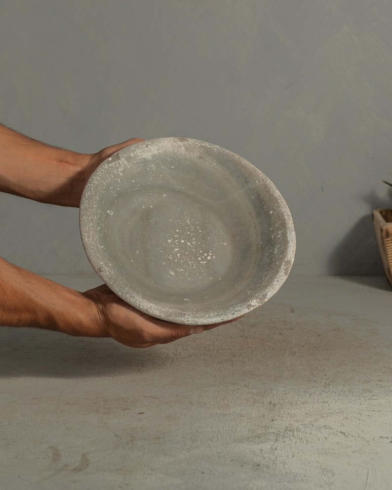 May include: A gray, round, stone bowl with a textured surface. The bowl is held by two hands. The bowl has a light gray interior and a darker gray rim. The bowl is likely used for serving food or as a decorative item.