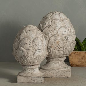 May include: Two decorative, weathered-looking artichoke sculptures in a neutral color palette. The sculptures have a textured surface and sit on square bases. The larger sculpture is approximately 12 inches tall, and the smaller one is about 9 inches tall.