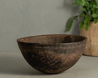 May include: A rustic, dark brown wooden bowl with a wide, shallow design. The bowl has visible wood grain and a textured surface, suggesting it is handmade. The bowl is empty and sits on a light gray surface, with a plant in the background.