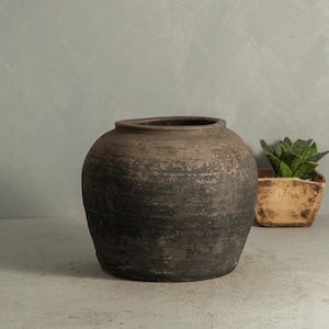 May include: A large, round, dark brown ceramic vase with a wide opening. The vase has a textured surface and a slightly uneven shape. A small, square wooden planter with a green plant is in the background.