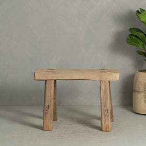 May include: A rectangular wooden stool with a light brown, weathered finish. The stool features four straight legs connected to a thick, flat seat with visible joinery details on the top surface, showcasing a natural, raw wood grain texture.