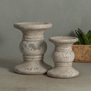 May include: Two weathered, off-white candle holders with a distressed finish. The holders have a classic, pedestal design with a wide base, a central bulbous section, and a flat top. The smaller holder is next to the larger one. A plant is in the background.