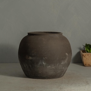 May include: A dark brown, round ceramic pot with a wide opening. The pot has a slightly textured surface and a weathered appearance, suggesting it may be vintage or handmade. The pot is likely used for decorative purposes or as a planter.
