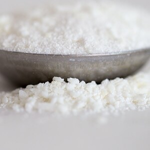 May include: A close-up of a metal measuring spoon filled with white powder. The powder is also scattered on the surface below the spoon.