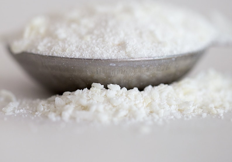 May include: A close-up of a metal measuring spoon filled with white powder. The powder is also scattered on the surface below the spoon.