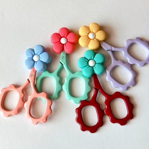 May include: A collection of colorful, flower-topped scissors in shades of pink, blue, red, yellow, green, and purple. The scissors have a scalloped edge design and are arranged on a white surface. These decorative scissors are ideal for crafting and sewing.