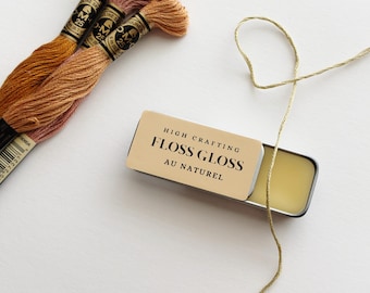 May include: A rectangular metal tin labeled High Crafting Floss Gloss Au Naturel containing a solid wax substance, accompanied by three skeins of embroidery thread in shades of orange and pink and a single strand of metallic gold thread.