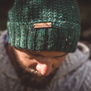 Op de afbeelding: Een groene gebreide beanie met een bruin leren label waarop "Sheena Knit" staat.