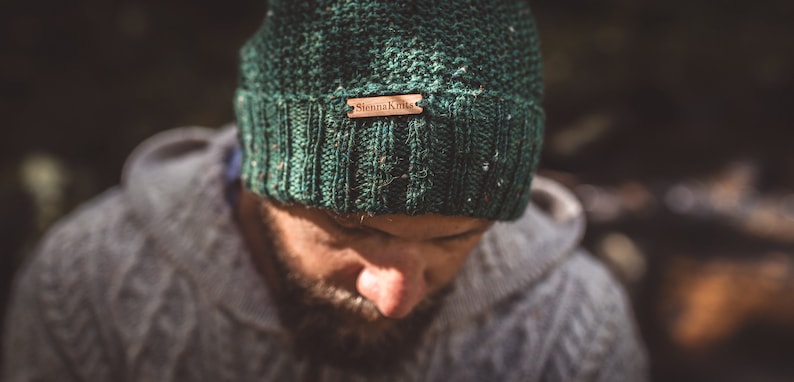 Op de afbeelding: Een groene gebreide beanie met een bruin leren label waarop "Sheena Knit" staat.