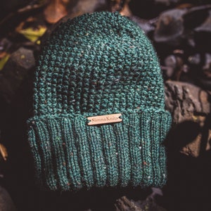 Op de afbeelding: Een groene gebreide beanie met een bruin leren label met de tekst "Sienna Knits".