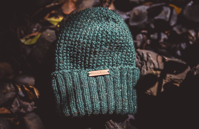 Op de afbeelding: Een groene gebreide beanie met een bruin leren label met de tekst "Sienna Knits".
