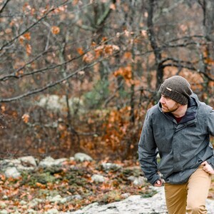 K&ouml;nnte beinhalten: Eine Person tr&auml;gt eine graue Jacke, eine braune M&uuml;tze und eine khakifarbene Hose und geht durch einen Wald. Die Jacke hat eine Kapuze und einen Rei&szlig;verschluss. Die M&uuml;tze ist dunkelbraun. Der Wald hat B&auml;ume mit orangefarbenen Bl&auml;ttern.