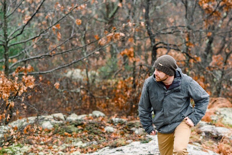 K&ouml;nnte beinhalten: Eine Person tr&auml;gt eine graue Jacke, eine braune M&uuml;tze und eine khakifarbene Hose und geht durch einen Wald. Die Jacke hat eine Kapuze und einen Rei&szlig;verschluss. Die M&uuml;tze ist dunkelbraun. Der Wald hat B&auml;ume mit orangefarbenen Bl&auml;ttern.