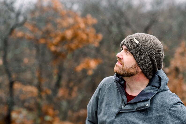 K&ouml;nnte beinhalten: Eine Person tr&auml;gt eine dunkelgraue M&uuml;tze mit einem kleinen Etikett, eine graue Jacke und ein rotes Hemd. Der Hintergrund zeigt verschwommenes Herbstlaub in Braun- und Oranget&ouml;nen.