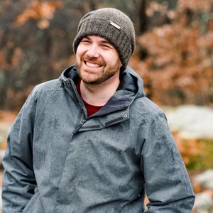 K&ouml;nnte beinhalten: Eine Person tr&auml;gt eine graue Kapuzenjacke und eine braune Strickm&uuml;tze. Die Jacke hat einen Rei&szlig;verschluss und einen dunkelgrauen Kragen. Die Person l&auml;chelt, ein rotes Hemd ist darunter sichtbar.