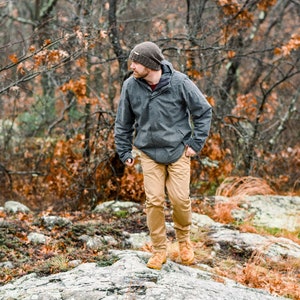 K&ouml;nnte beinhalten: Eine Person mit grauer Jacke, beigefarbener Hose und braunen Stiefeln geht auf einer felsigen Oberfl&auml;che. Sie tr&auml;gt auch eine Strickm&uuml;tze. Der Hintergrund zeigt B&auml;ume mit herbstlich gef&auml;rbten Bl&auml;ttern.