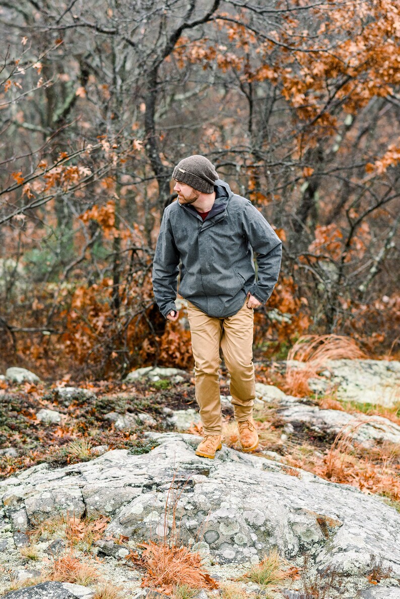 K&ouml;nnte beinhalten: Eine Person mit grauer Jacke, beigefarbener Hose und braunen Stiefeln geht auf einer felsigen Oberfl&auml;che. Sie tr&auml;gt auch eine Strickm&uuml;tze. Der Hintergrund zeigt B&auml;ume mit herbstlich gef&auml;rbten Bl&auml;ttern.