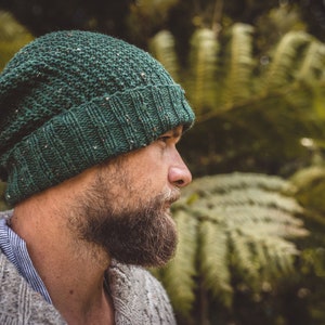 Op de afbeelding: Een man draagt ​​een groene gebreide beanie met een textuurpatroon. Hij heeft een baard en kijkt naar de zijkant. De achtergrond is wazig en lijkt een bos te zijn.