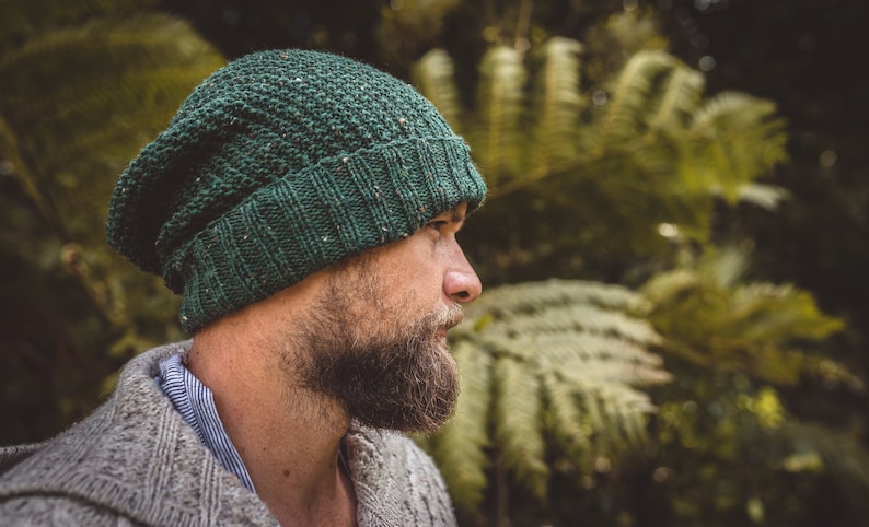 Op de afbeelding: Een man draagt ​​een groene gebreide beanie met een textuurpatroon. Hij heeft een baard en kijkt naar de zijkant. De achtergrond is wazig en lijkt een bos te zijn.